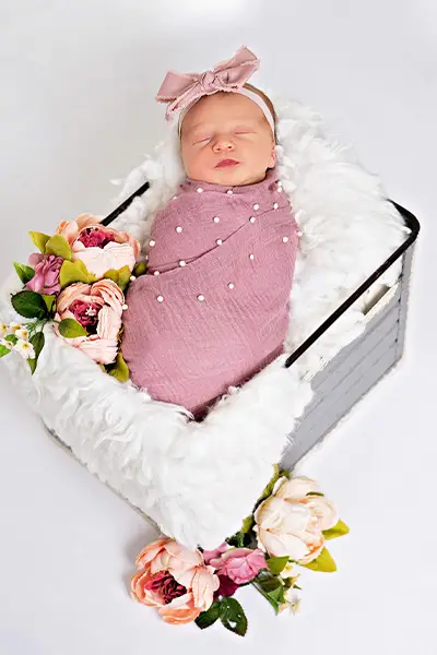 Newborn baby girl wrapped in a pink, pearl-studded blanket with a matching headband, lying in a white basket decorated with flowers, photographed by Dia Kiss, a Chicago suburbs photographer.