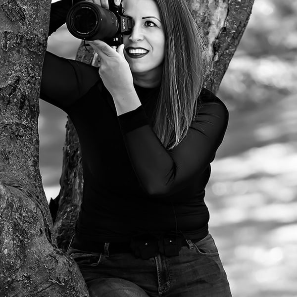 A black-and-white photo of Diana Kiss, a photographer in Chicago, leaning against a tree with her camera raised, wearing a black sheer top and jeans, and smiling while capturing a shot in a natural outdoor setting.