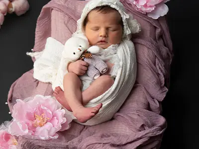 Newborn photoshoot featuring a baby girl sleeping peacefully, wrapped in a white blanket with a bonnet, holding a small stuffed animal, surrounded by pink flowers on a soft background.