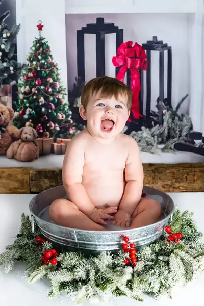 Family photoshoot featuring a 1-2 year old child sitting in a festive metal tub surrounded by holiday decorations, including Christmas trees and teddy bears.