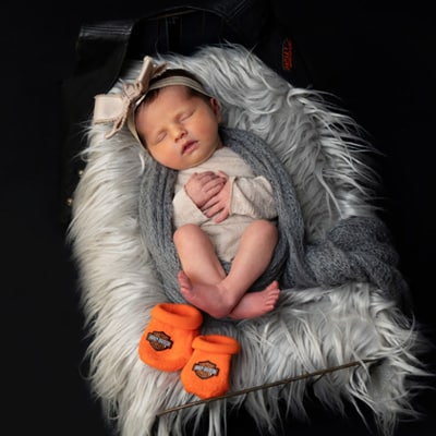 A newborn baby sleeping on a white faux fur blanket, wrapped in a gray knit scarf, with Harley Davidson-branded orange booties at their feet. The baby is wearing a beige headband with a bow, and a black background is positioned behind. Captured by a Chicago suburbs newborn photographer.