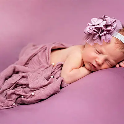 Newborn baby girl wrapped in a soft purple blanket, adorned with a matching flower headband, peacefully sleeping on a purple backdrop, taken by a Chicago suburbs newborn photographer.