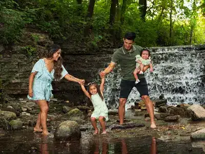 Outdoor family photoshoot by a waterfall, featuring a joyful family with parents holding hands with their young daughter while holding an infant, surrounded by lush greenery.