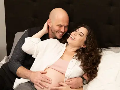 Maternity photoshoot featuring a happy couple lounging on a bed, with the man embracing the woman’s pregnant belly as they both smile and laugh together.