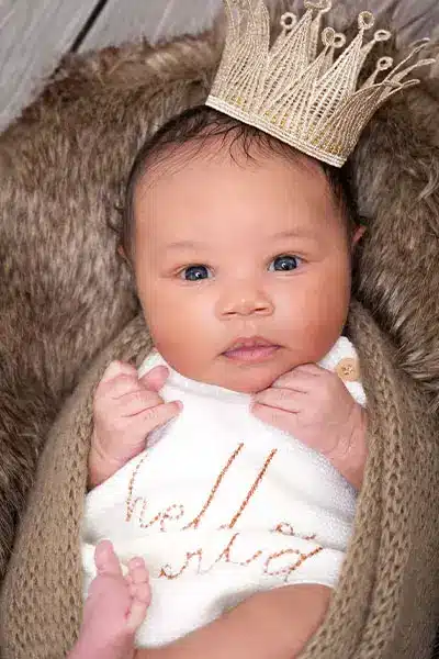 Newborn baby girl wrapped in a soft brown blanket, wearing a "Hello World" outfit and a gold crown, photographed by a Chicago suburbs photographer.