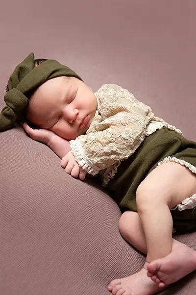 Newborn baby girl peacefully sleeping on a soft pink backdrop, wearing a green lace outfit and matching headband, photographed by a Chicago suburbs photographer.