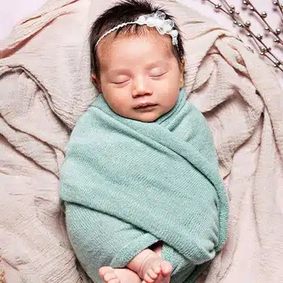 Newborn baby girl wrapped in a mint-colored blanket with a delicate headband, sleeping on a soft, textured background with natural accents around her.