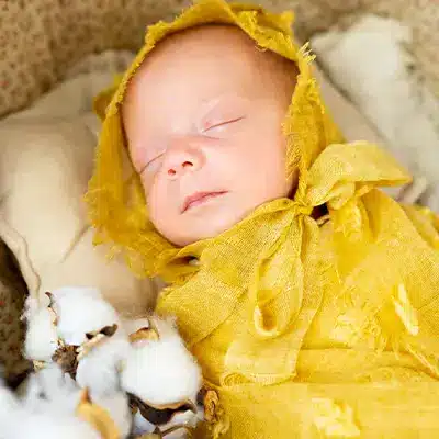 Newborn baby wrapped in a bright yellow fabric with a matching bonnet, resting peacefully with cotton plant decorations nearby.