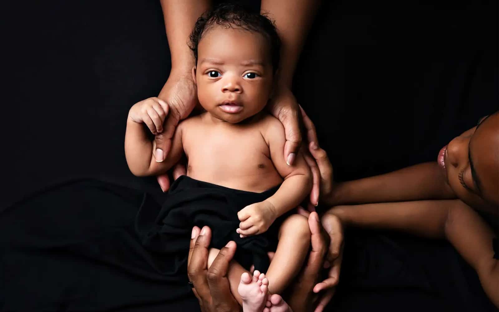 A newborn baby lying on a black background, surrounded and gently held by multiple hands, symbolizing family unity and care. Photo by Dia Kiss in Chicago suburbs.