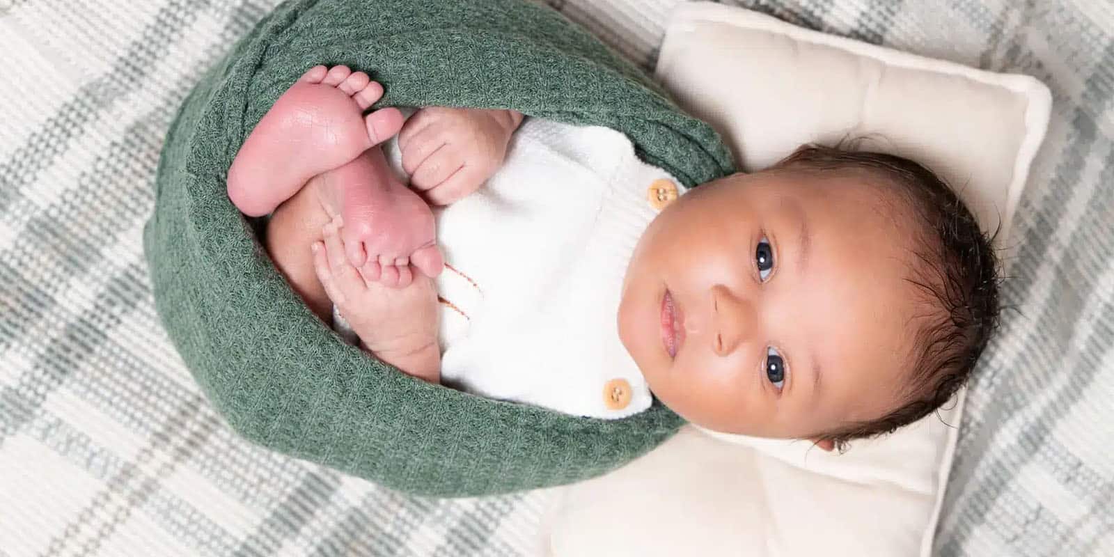 Newborn baby wrapped in a green blanket, lying on a plaid-patterned blanket, looking up at the camera.