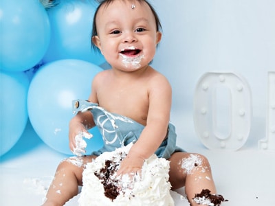 A baby smiling during a cake smash photoshoot in a Chicago suburbs studio, sitting on the floor with blue balloons in the background and frosting smeared on their face and hands.