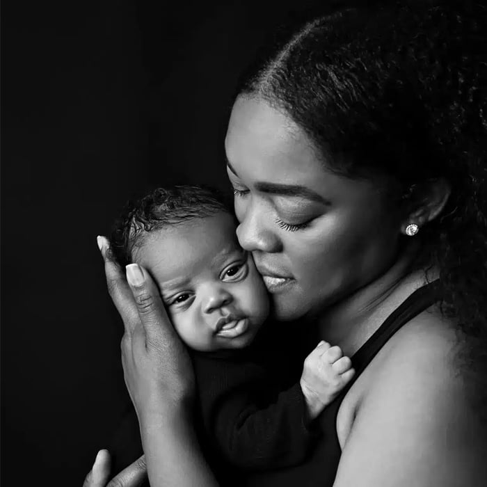 Black and white portrait of a mother holding her newborn close, captured by Chicago newborn photographer Dia Kiss. The mother cradles the baby with a gentle expression, conveying warmth and tenderness against a dark background.