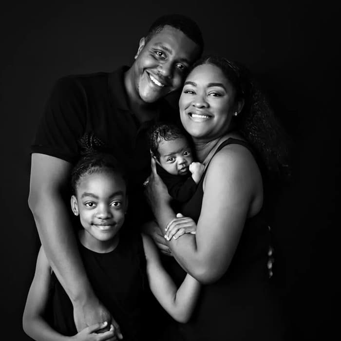 Black and white family portrait featuring a father, mother, young daughter, and newborn baby, all smiling warmly, captured by Crystal Lake, IL newborn photographer Dia Kiss. The family is dressed in black, embracing each other closely, creating a loving and connected image.