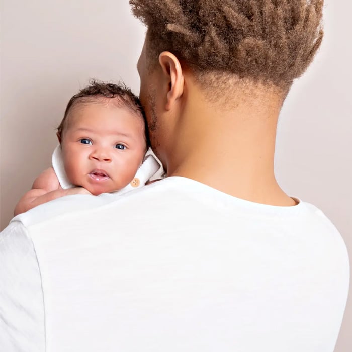 Newborn baby resting on their father’s shoulder, looking alert and curious, captured by Crystal Lake, IL newborn photographer Dia Kiss. The father faces away, wearing a white shirt, while the baby gazes directly at the camera against a soft, neutral background.