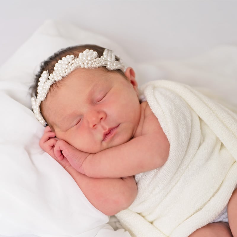 Infant photoshoot of a newborn baby sleeping wrapped in a soft white blanket, wearing a pearl headband on a white background.