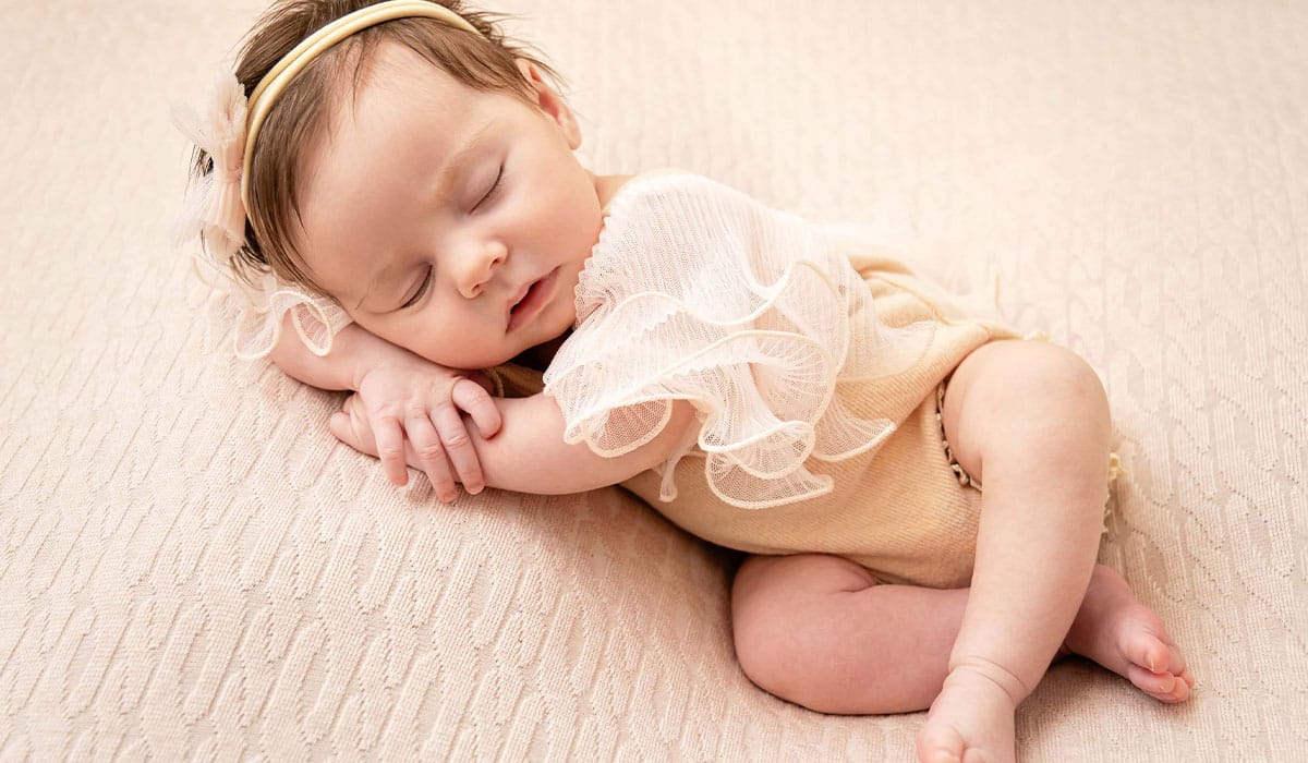 A newborn baby girl peacefully sleeping on a textured blanket, wearing a delicate headband and ruffled outfit.