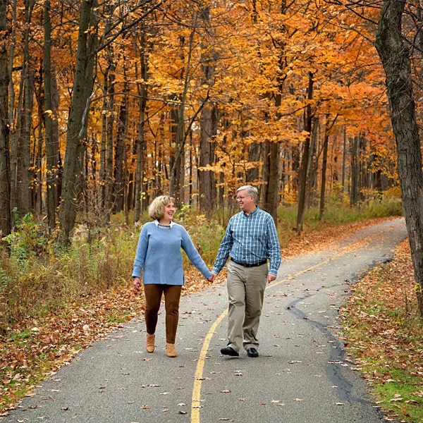 A couple holding hands and walking on a tree-lined path surrounded by vibrant orange and yellow autumn foliage.