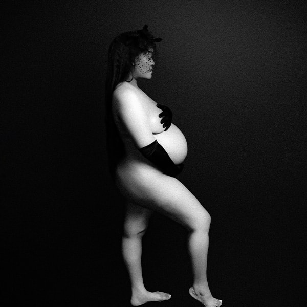 An artistic black and white image of a pregnant woman posing against a dark background, wearing long gloves and a veiled headpiece, emphasizing silhouette and form.