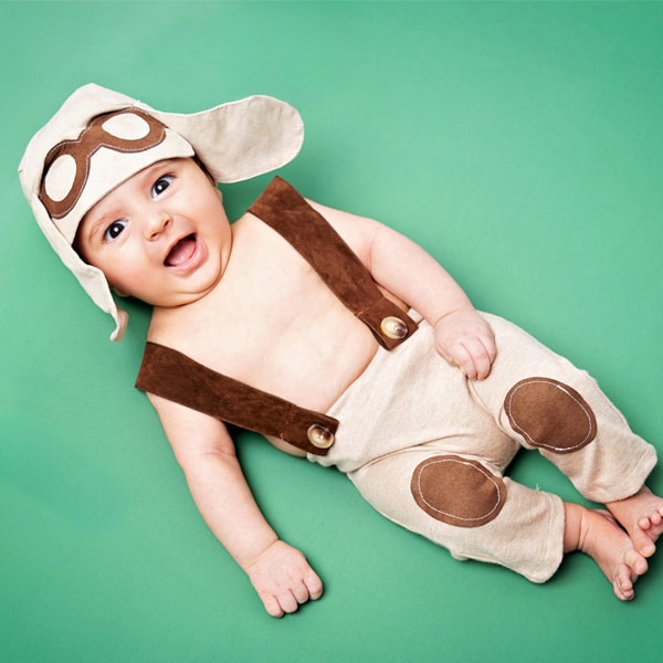 A smiling baby wearing an aviator costume, lying on a green background.