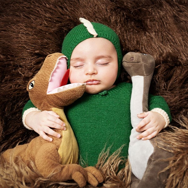 A baby dressed in a green dinosaur-themed outfit, sleeping while hugging two plush dinosaur toys on a soft brown textured surface.