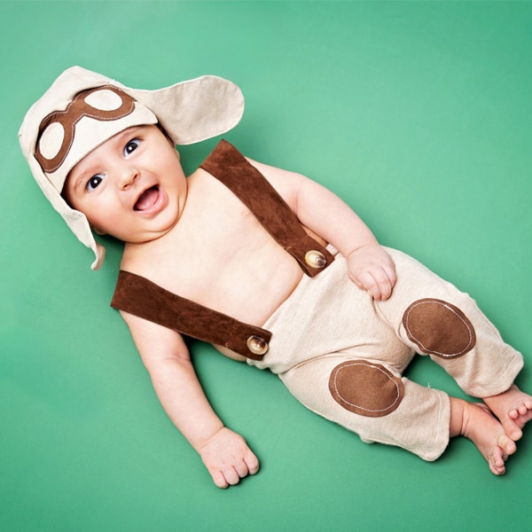 A smiling baby dressed as a little aviator, wearing suspenders and a pilot hat, lying on a green backdrop.