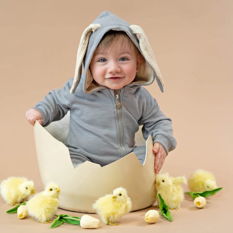 A baby in a gray bunny hoodie sitting inside a large egg shell prop, surrounded by yellow plush chicks and tulips on a pastel background.