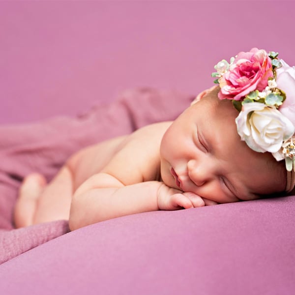 A sleeping newborn baby on a purple background, wearing a colorful floral headband and resting on a soft blanket.