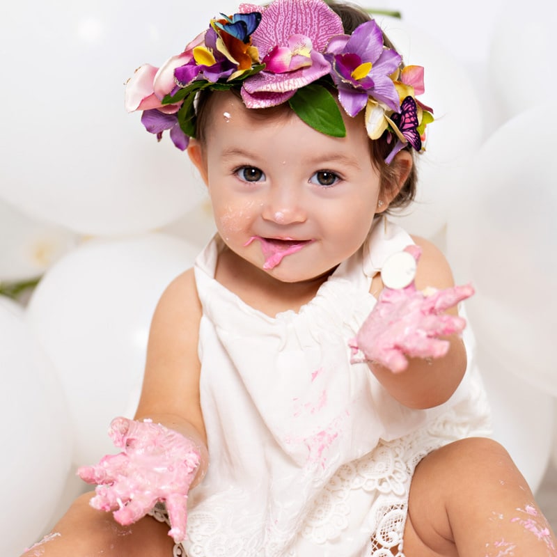 A baby girl wearing a colorful flower crown, enjoying a cake smash with pink frosting on her hands and face, surrounded by white balloons.