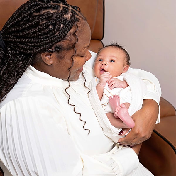 A baby in a white outfit being held by a smiling mother with braided hair, seated in a brown chair.