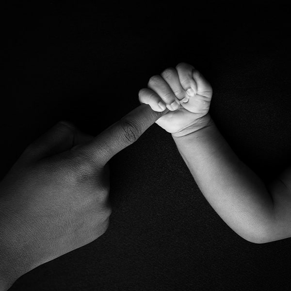 A black-and-white close-up of a newborn's hand gripping an adult's finger against a dark background.