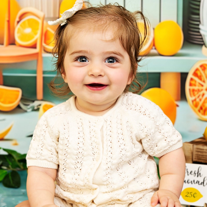 A smiling baby in a white outfit sitting in front of a lemonade stand backdrop with oranges and lemons, holding a wooden toy.