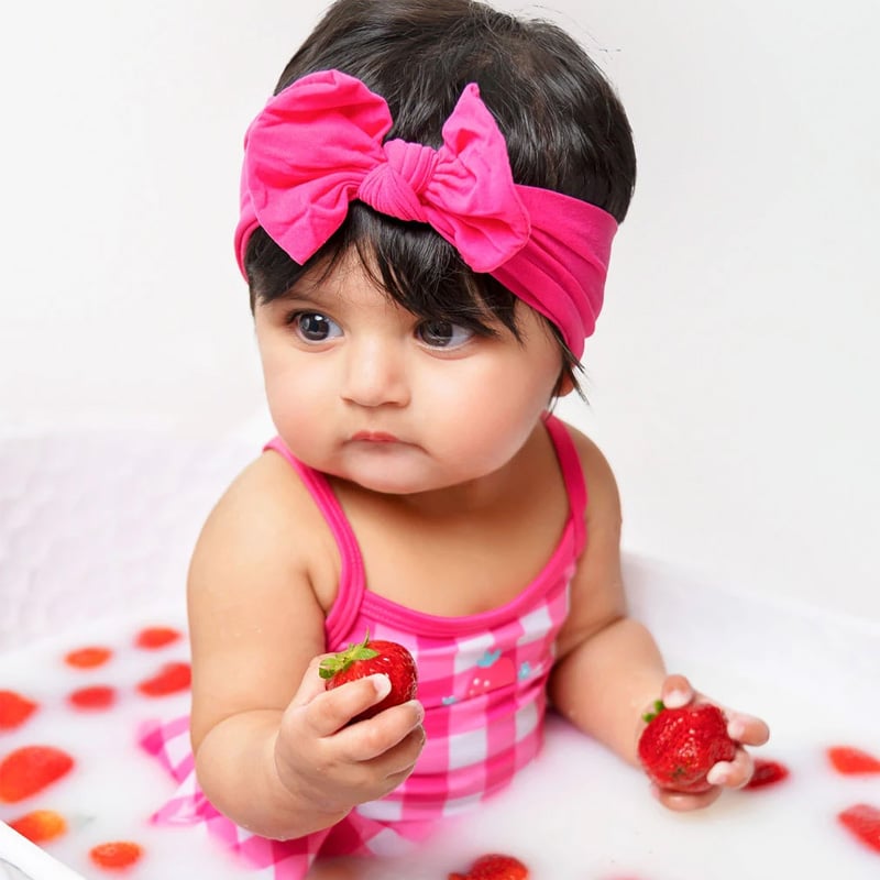 A baby in a pink outfit with a matching headband sitting in a milk bath surrounded by fresh strawberries.