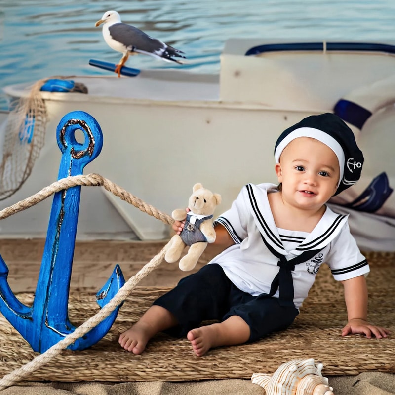 A baby dressed in a sailor outfit holding a teddy bear, seated next to a blue anchor with a nautical-themed background.