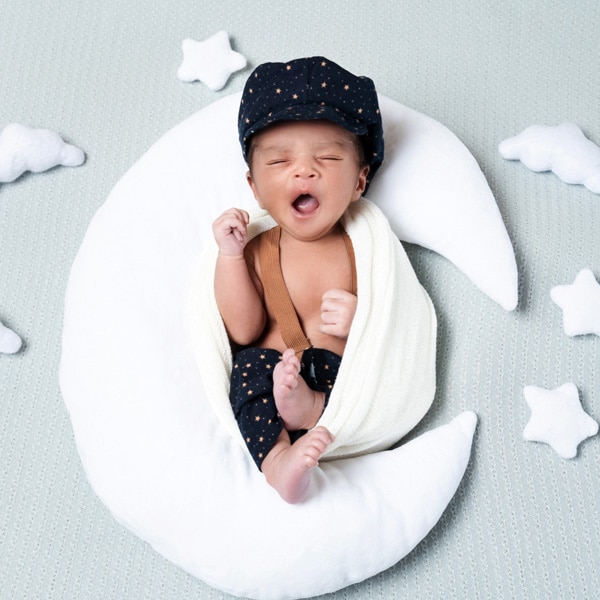 A yawning baby dressed in a star-patterned hat and outfit, lying on a crescent moon-shaped pillow surrounded by plush stars and clouds.