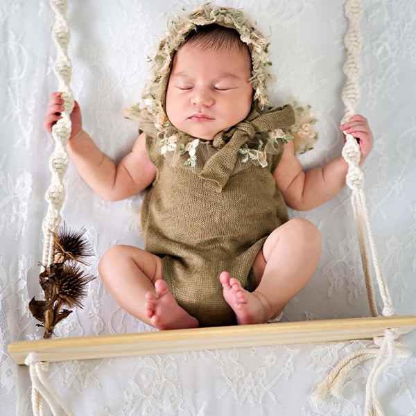 A baby dressed in a floral bonnet and green outfit, peacefully sleeping on a swing against a white lace background.