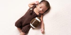 A baby dressed in a brown knitted outfit, peacefully sleeping while holding a small plush football.