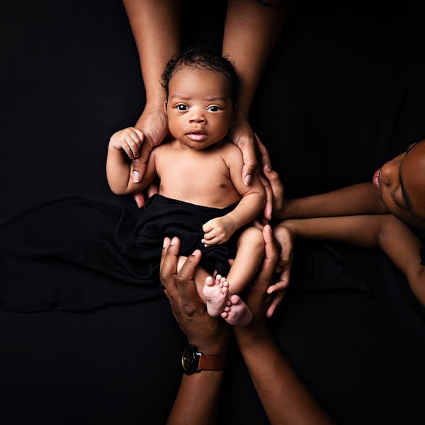 A baby lying on a black background, surrounded and supported by adult hands, looking alert and curious.