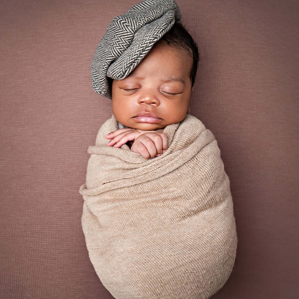 A baby swaddled in beige fabric, wearing a herringbone-patterned cap, sleeping peacefully on a textured brown background.