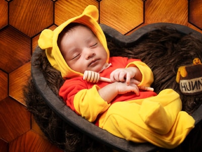 A baby dressed in a Winnie the Pooh-inspired costume, sleeping in a basket while holding a wooden honey dipper, with a small 'Hunny' pot beside them against a honeycomb wooden background.