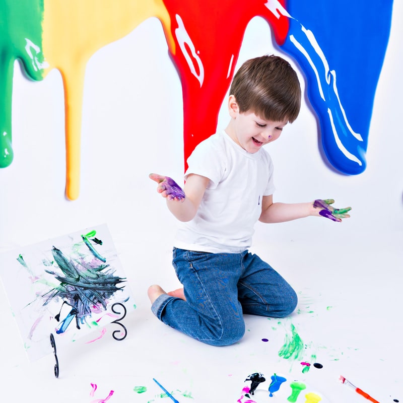 A young boy in a white shirt and jeans playing with colorful paints on a white canvas, with vibrant paint splashes on the background.