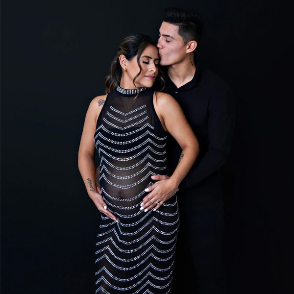 A couple embracing for a maternity portrait, with the pregnant woman wearing a black gown adorned with decorative patterns and her partner kissing her forehead against a black background.