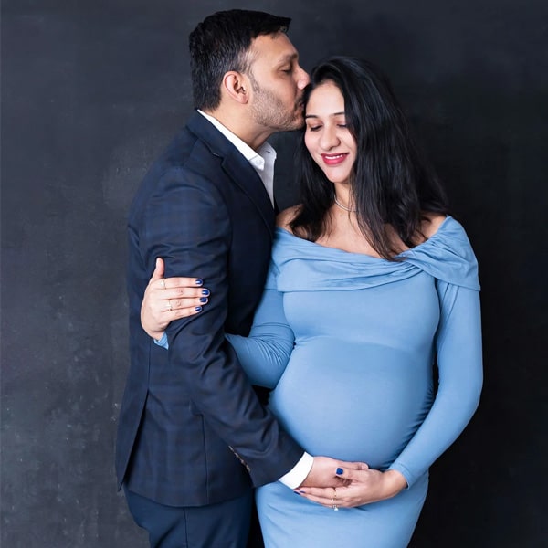 A pregnant woman in a fitted blue dress smiling as her partner kisses her forehead, both cradling her baby bump against a dark textured background.