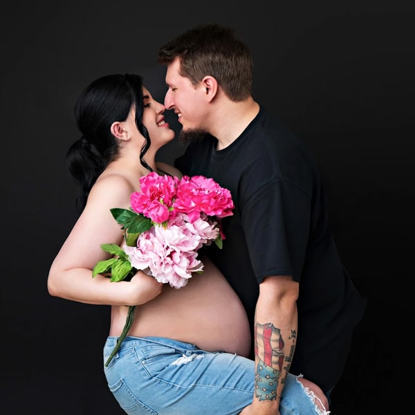 A pregnant woman holding a bouquet of pink flowers while smiling and standing close to her partner, who leans toward her affectionately, against a black background.