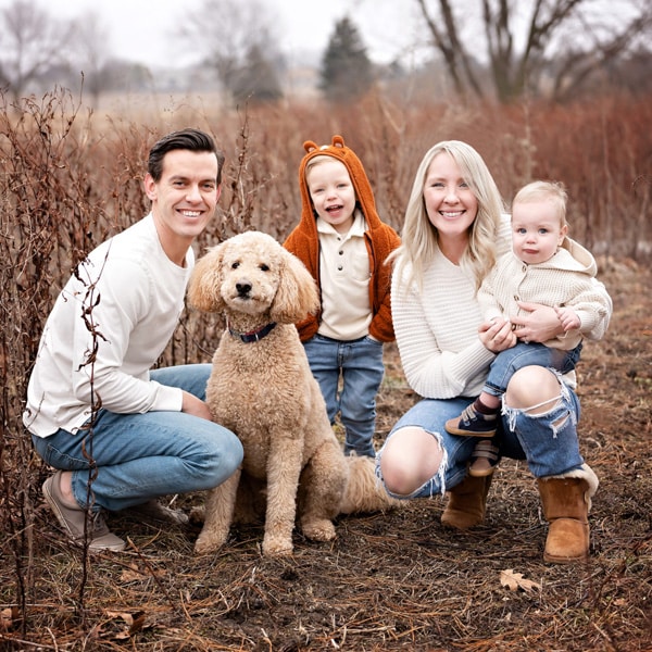 A family of four with a golden doodle dog posing outdoors in a fall field, surrounded by dried vegetation and trees in the background.