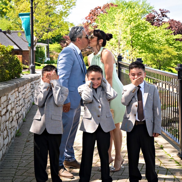 A couple kissing outdoors on a sunny day, while three boys in matching suits humorously cover their eyes, ears, and mouth in a playful pose.