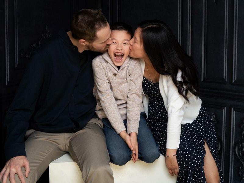 A joyful family moment with parents kissing their laughing child on the cheeks, set against a dark, elegant backdrop.