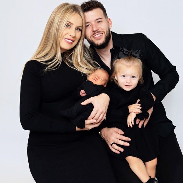 A smiling family dressed in black, with a mother holding a newborn baby and a young child sitting on their father's lap.