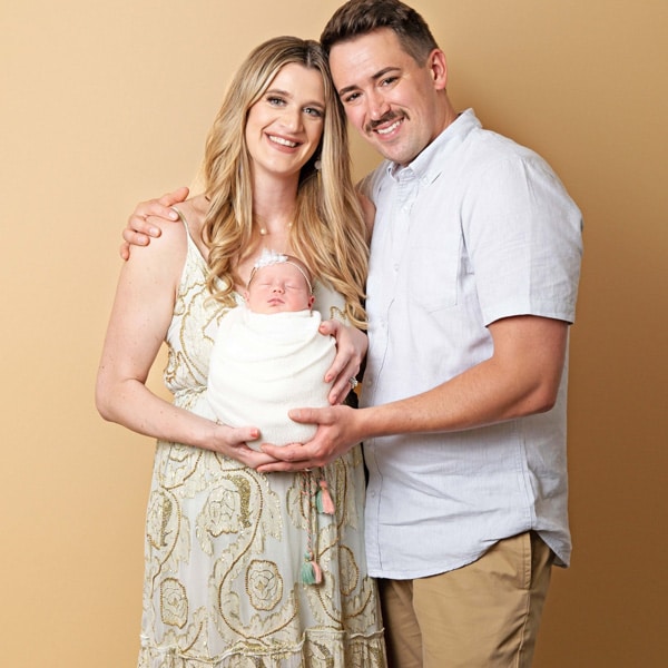 A smiling couple holding their newborn baby swaddled in white, standing against a soft beige background.