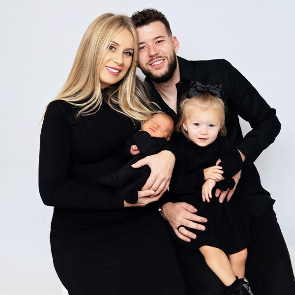 A family portrait featuring a mother, father, toddler, and newborn, all dressed in black, posing together against a white background.