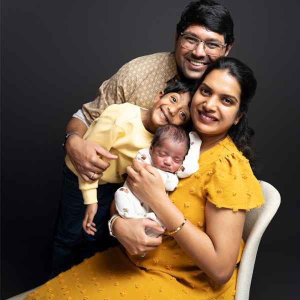 A family portrait featuring a mother in a yellow dress holding a sleeping newborn, with a father and older child smiling in the background.